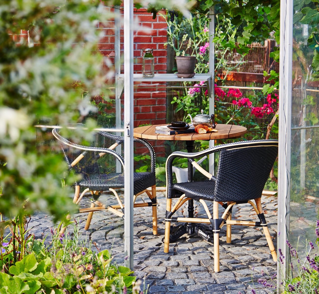 Esta es una imagen de estilo de vida de Madeleine silla de comedor en black ArtFibre skin on rattan frame. Invernadero con suelo de adoquines y plantas.