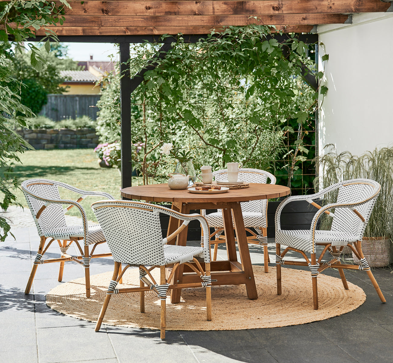 Esta es una imagen de estilo de vida de Madeleine silla de comedor en white w cappuccino dot ArtFibre skin on rattan frame. Pérgola de jardín con alfombra de yute.