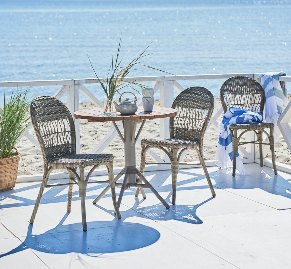 Esta es una imagen de estilo de vida de Ofelia Exterior Chair i antique grey artfibre aluminium. En una terraza junto al mar con barandilla blanca y toalla.