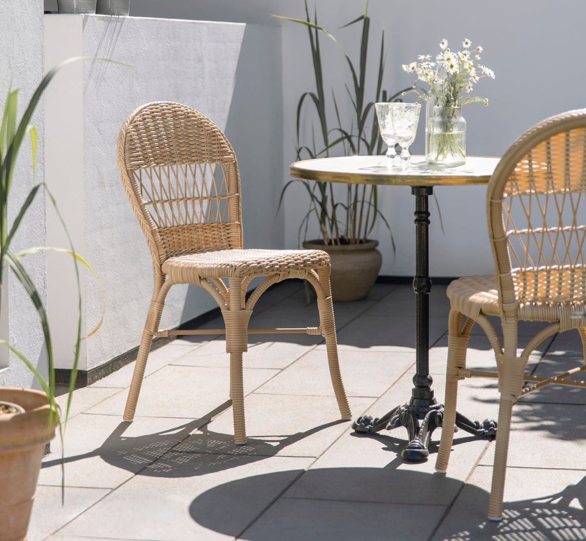Esta es una imagen de estilo de vida de Ofelia Exterior Chair i natural artfibre aluminium. En un patio soleado con margaritas y mesa de bistró.