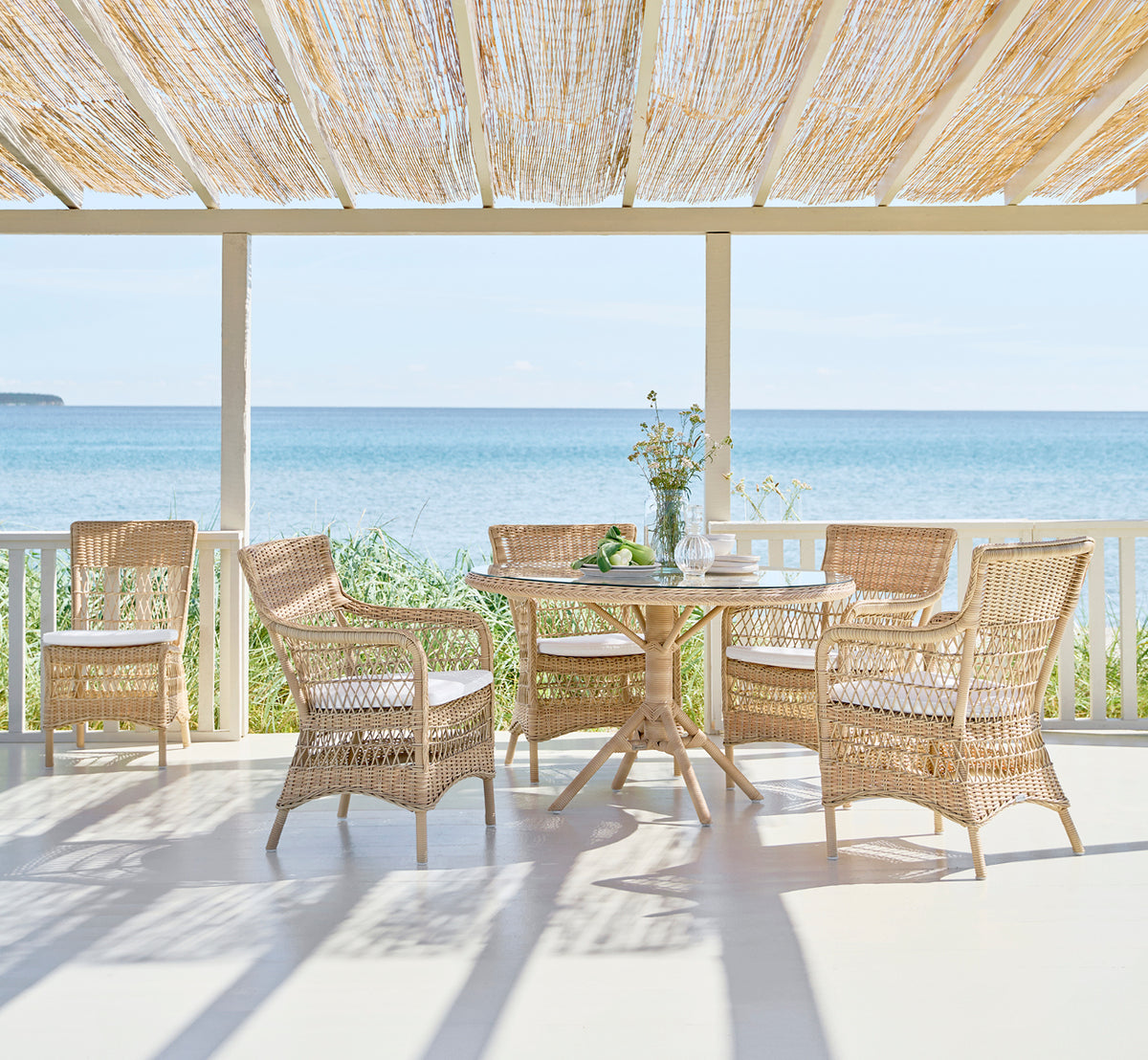 Esta es una imagen de estilo de vida de Marie Exterior Side Chair i natural artfibre aluminium. Alrededor de una mesa en una terraza sombreada junto al mar.