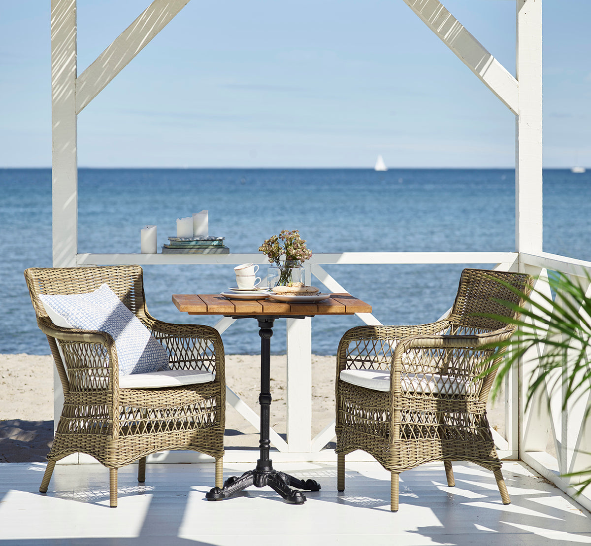 Esta es una imagen de estilo de vida de Marie Exterior Armchair i antique grey artfibre aluminium. Junto a una mesa de teca en un pabellón de playa.