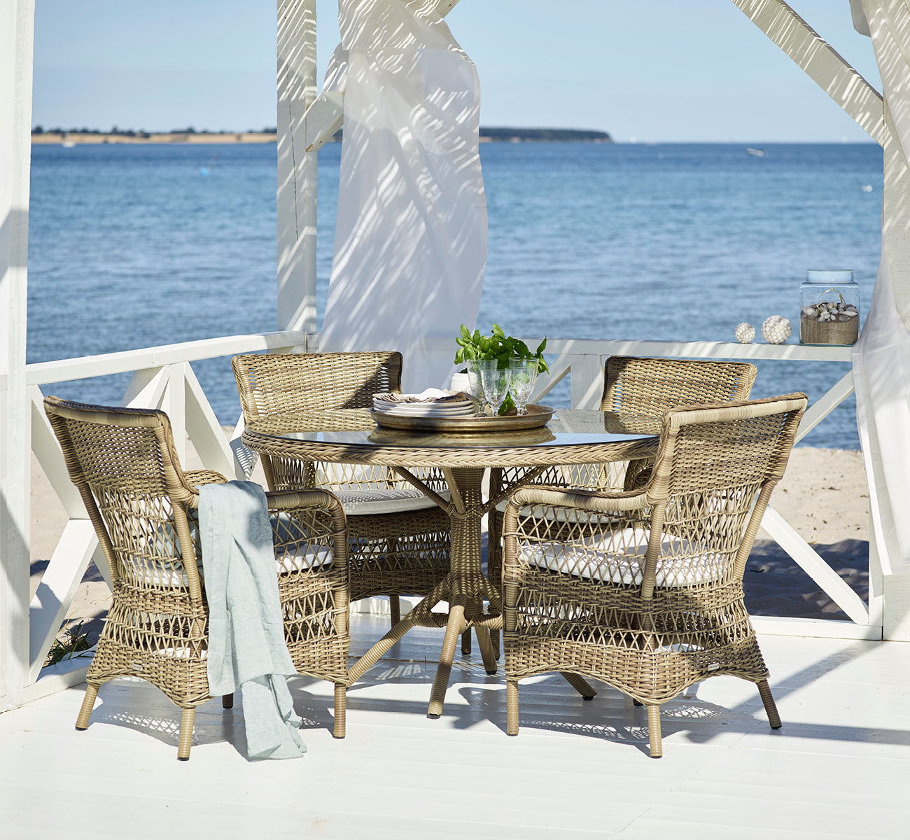 Esta es una imagen de estilo de vida de Marie Exterior Armchair i antique grey artfibre aluminium. En un pabellón cubierto junto al mar con el océano.