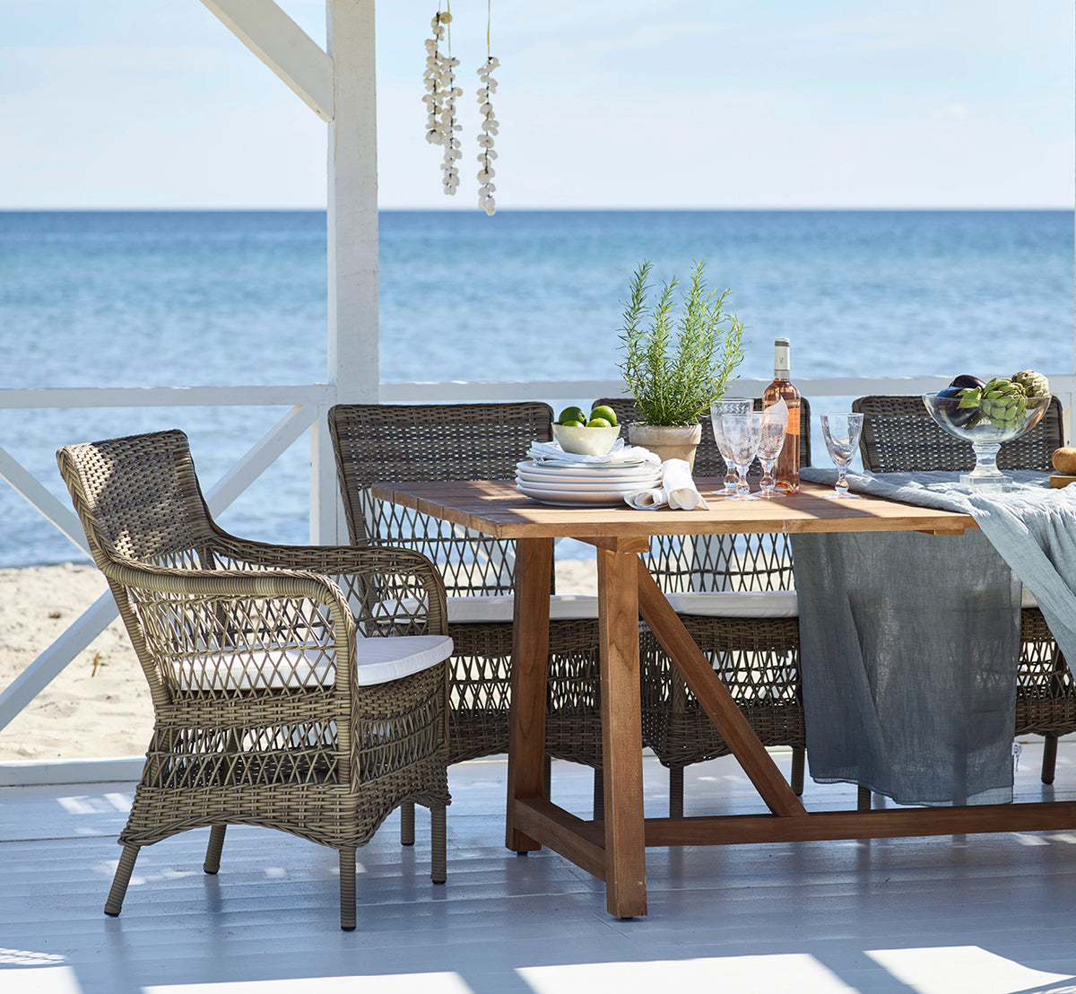Esta es una imagen de estilo de vida de Marie Exterior Armchair i antique grey artfibre aluminium. Alrededor de una mesa de teca en un pabellón de playa.