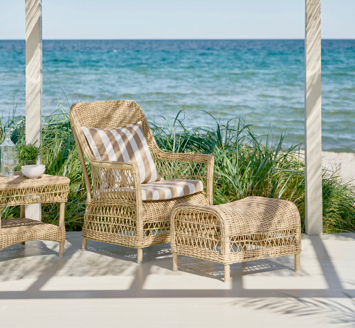 Esta es una imagen de estilo de vida de Dawn reposapiés en natural ArtFibre Aluminium, en una terraza costera con vistas al mar, gramíneas de playa y.