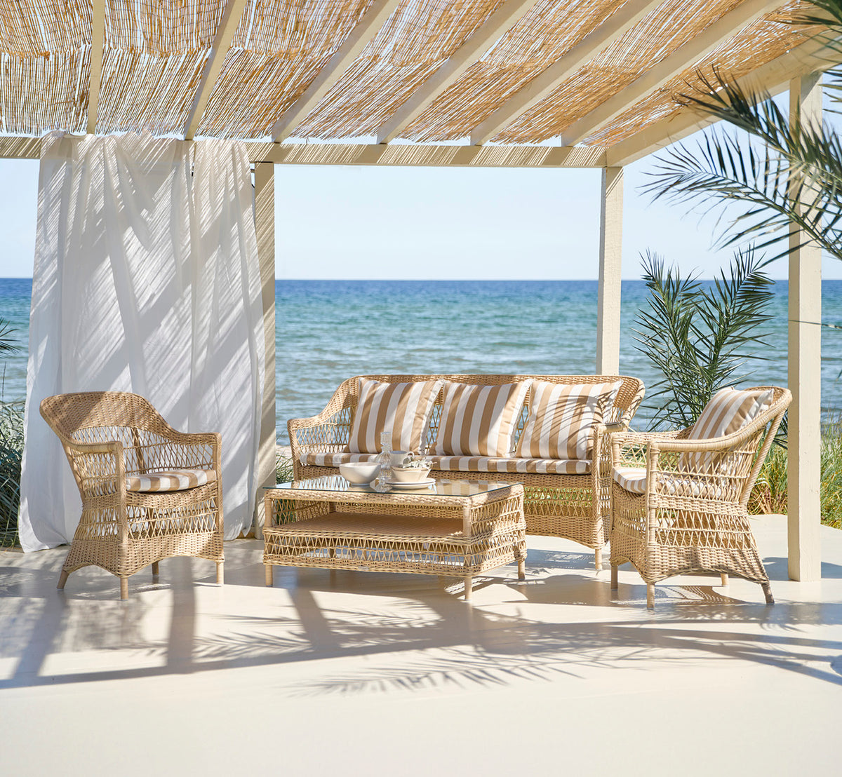 Esta es una imagen de estilo de vida de Charlot sofá de 3 plazas en natural ArtFibre Aluminium, en una terraza costera con techo de caña y vistas al.