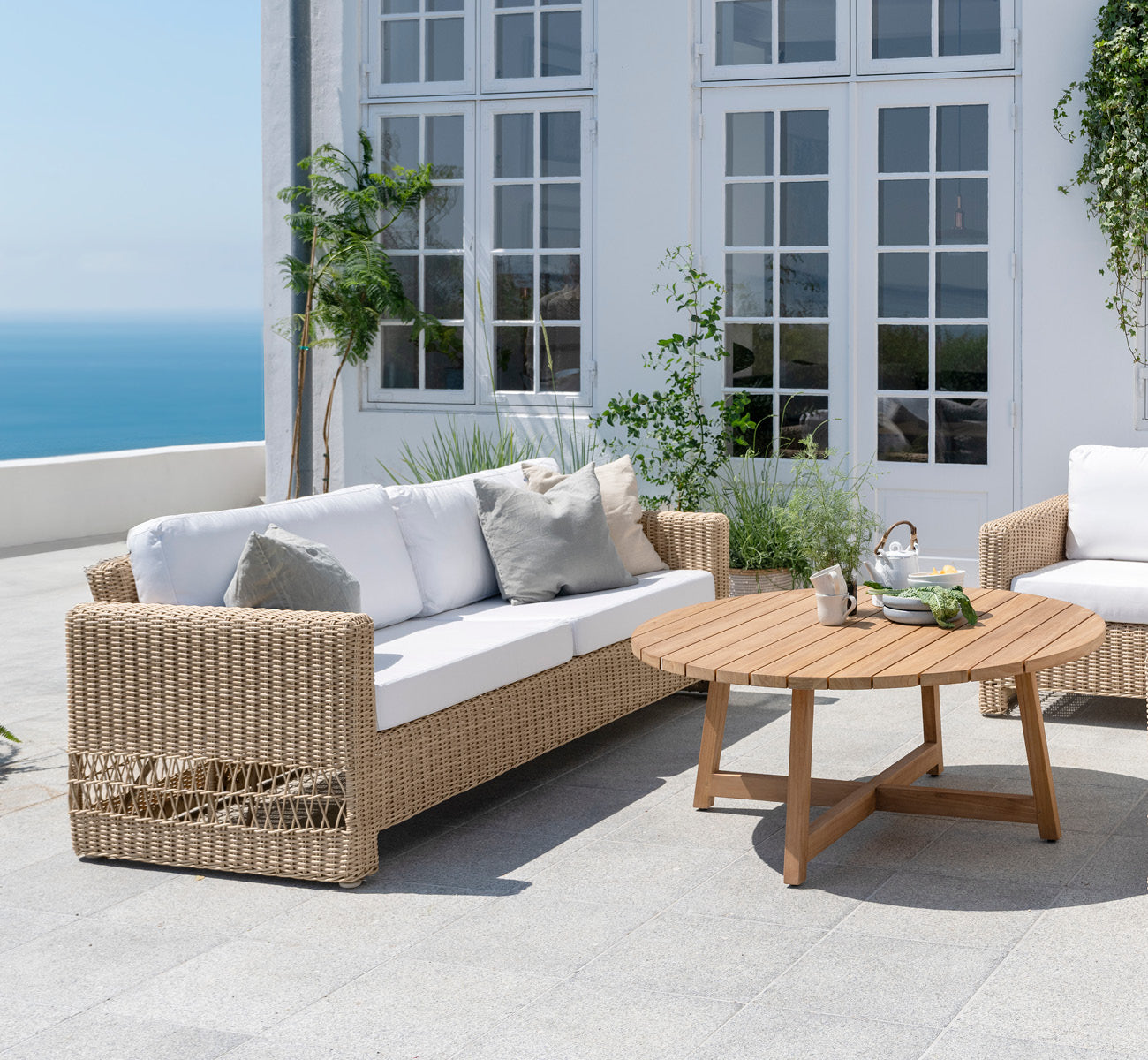 Esta es una imagen de estilo de vida de Carrie sofá en natural ArtFibre Aluminium, en una terraza costera con cubierta de madera blanca, vistas al.