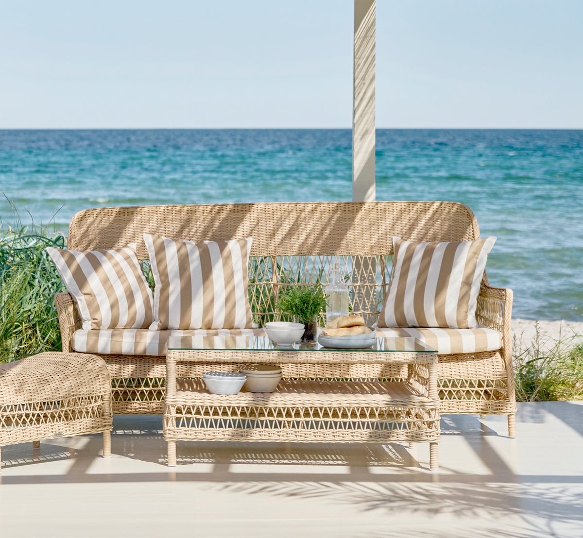 Esta es una imagen de estilo de vida de Dawn sofá en natural ArtFibre Aluminium, en una terraza costera con cubierta de madera blanca y vistas al mar.