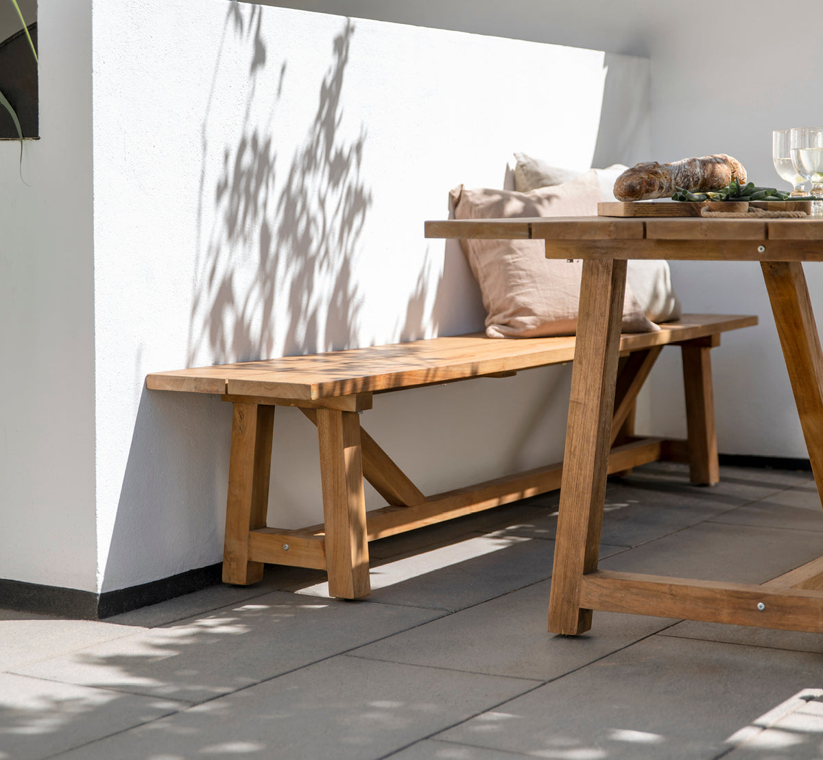 Esta es una imagen de estilo de vida de George banco exterior en outdoor Teak, en un patio exterior con suelo de baldosas grises y pared encalada.