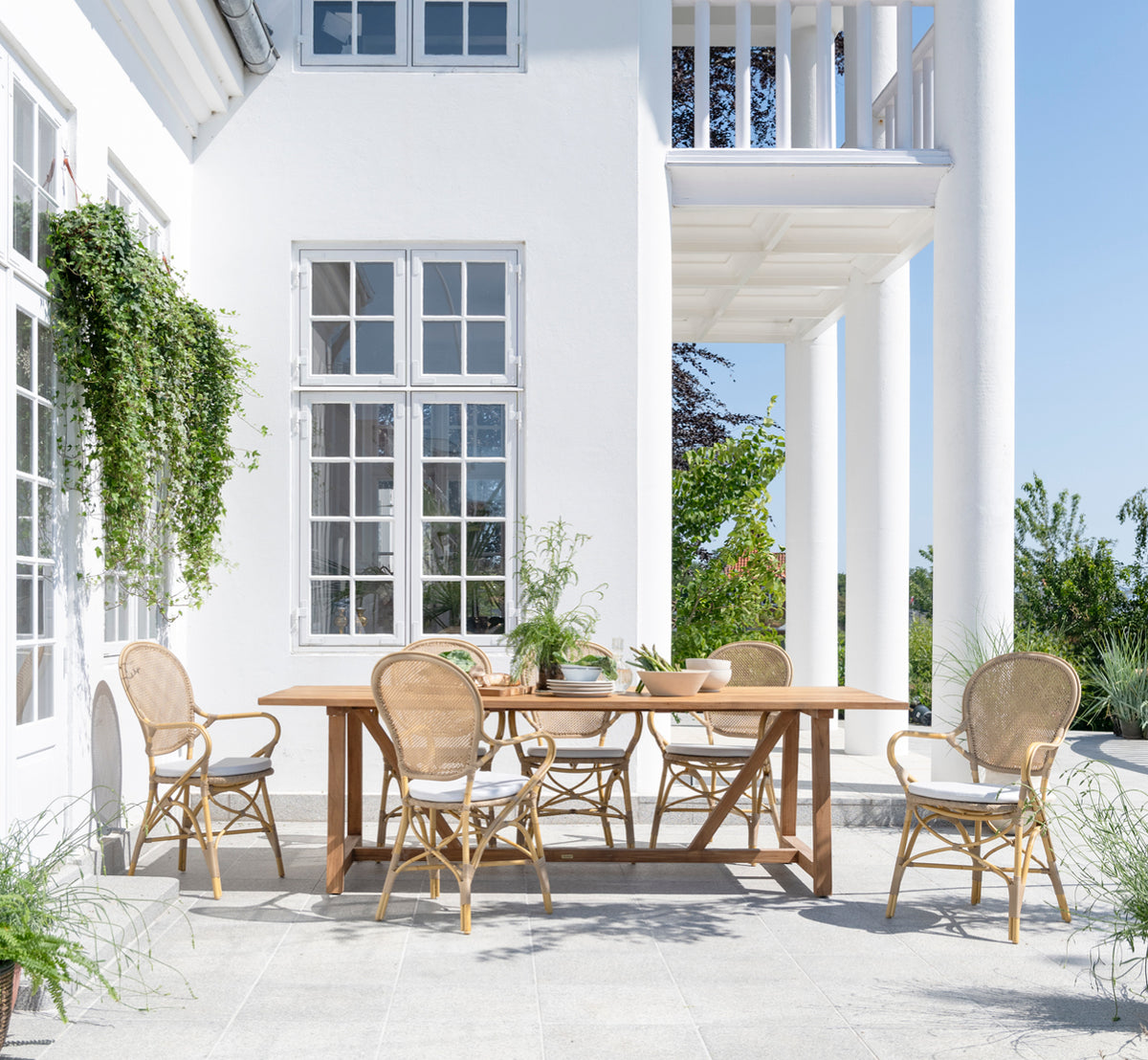 Esta es una imagen de estilo de vida de George mesa exterior 240 en outdoor Teak, en un patio de jardín exterior con fachada blanca y vegetación.