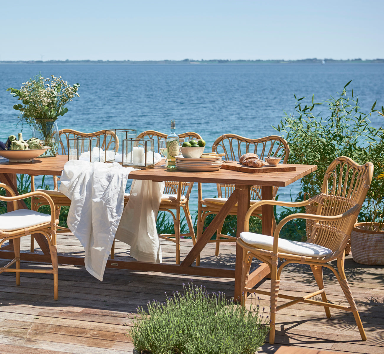 Esta es una imagen de estilo de vida de George mesa exterior 240 en outdoor Teak, en un patio de jardín exterior con fachada blanca y vegetación.