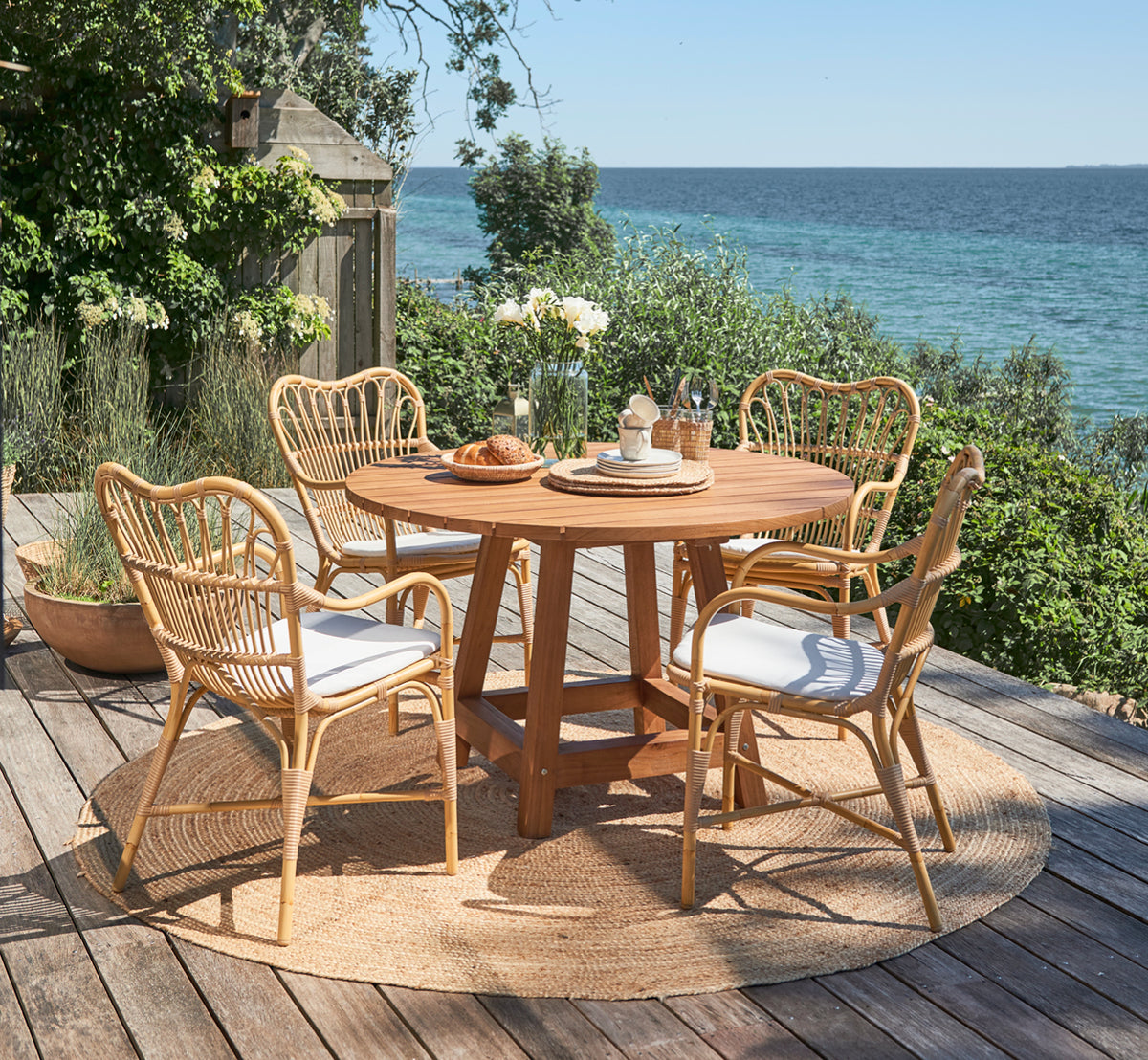 Esta es una imagen de estilo de vida de George mesa exterior Ø120 en outdoor Teak, en un patio de jardín exterior con fachada blanca y vegetación.
