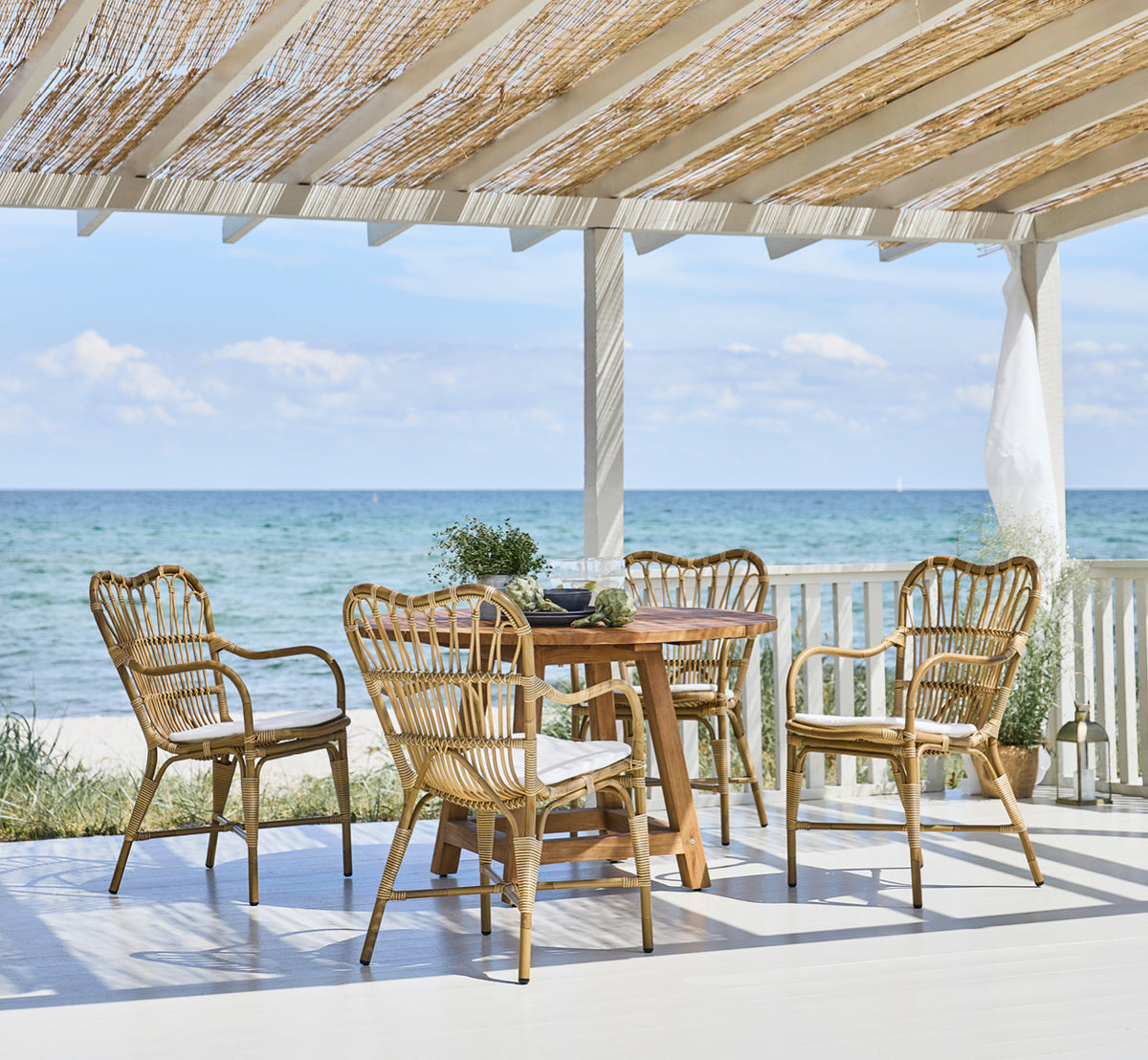 Esta es una imagen de estilo de vida de George mesa exterior Ø120 en outdoor Teak, en un patio de jardín exterior con fachada blanca y vegetación.