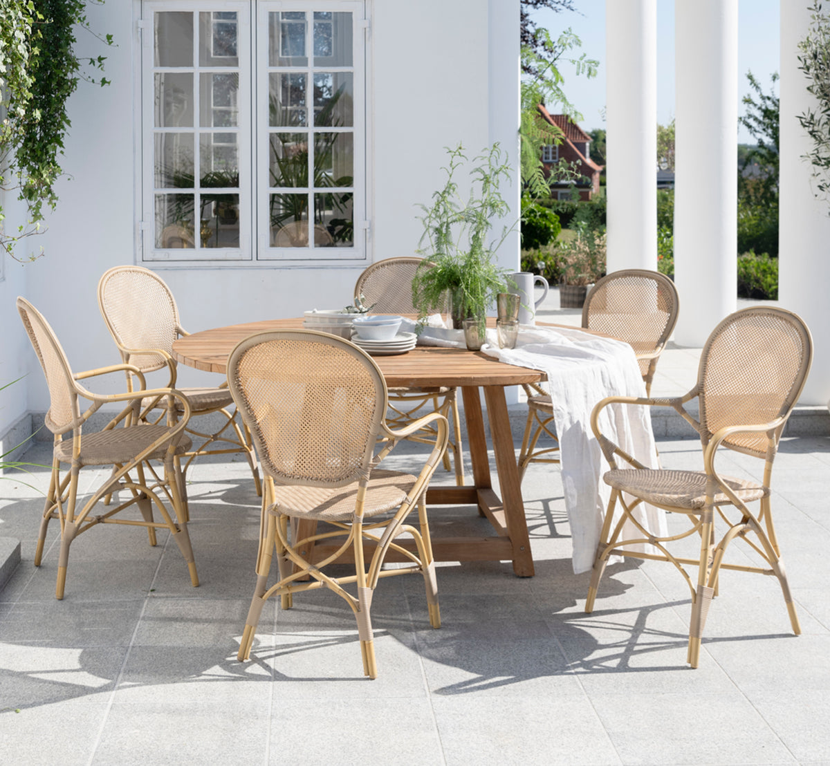 Esta es una imagen de estilo de vida de George mesa exterior Ø160 en outdoor Teak, en un patio de jardín exterior con fachada blanca y vegetación.