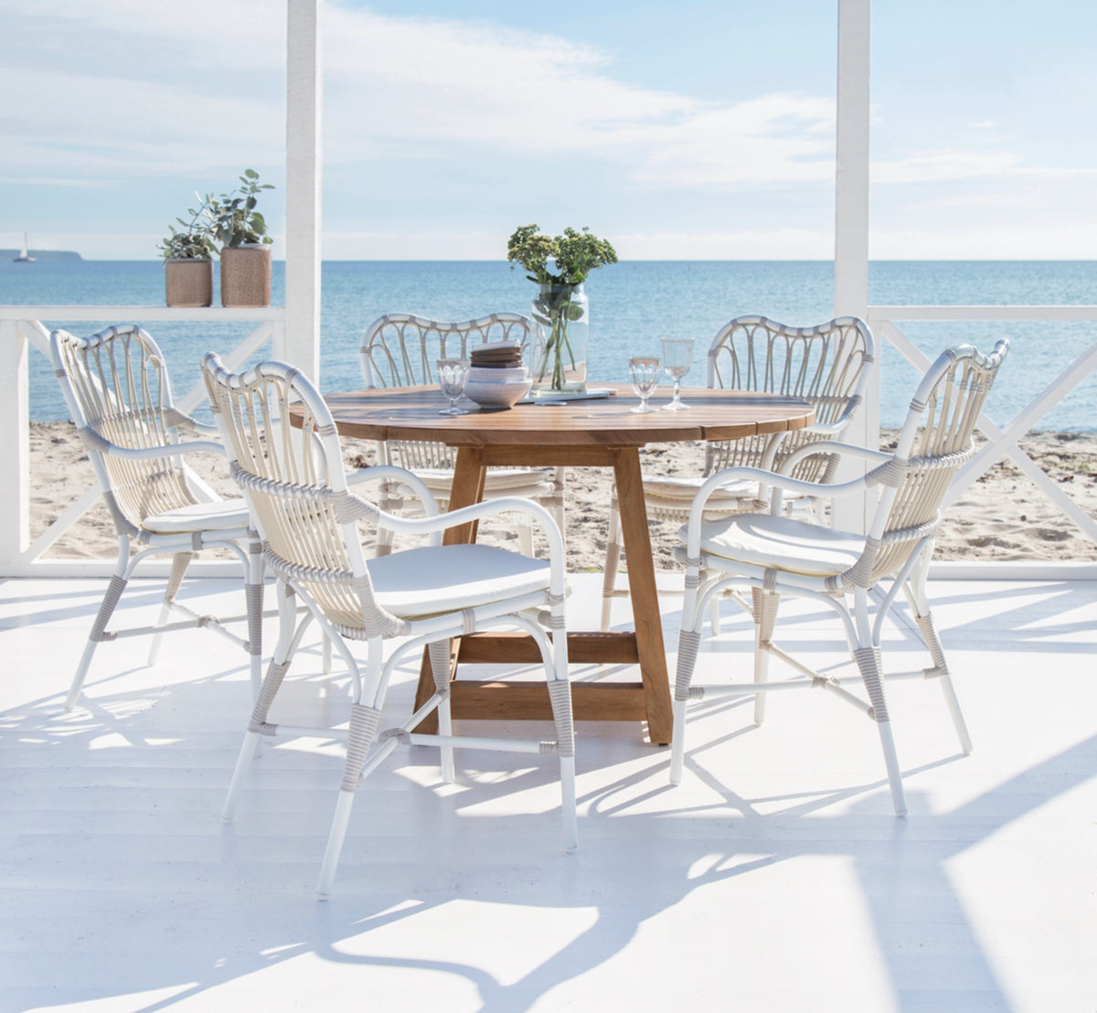 Esta es una imagen de estilo de vida de George mesa exterior Ø160 en outdoor Teak, en un patio de jardín exterior con fachada blanca y vegetación.