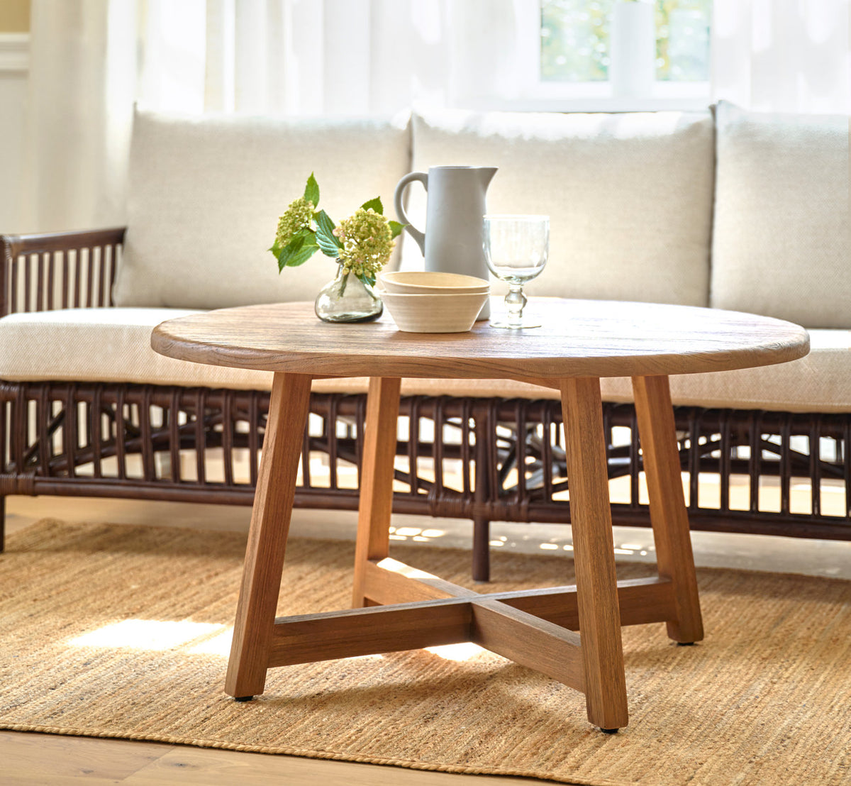 Esta es una imagen de estilo de vida de Aksel mesa de centro Ø100 cm en teak indoor. Delante de un sofá de ratán sobre una alfombra de yute.