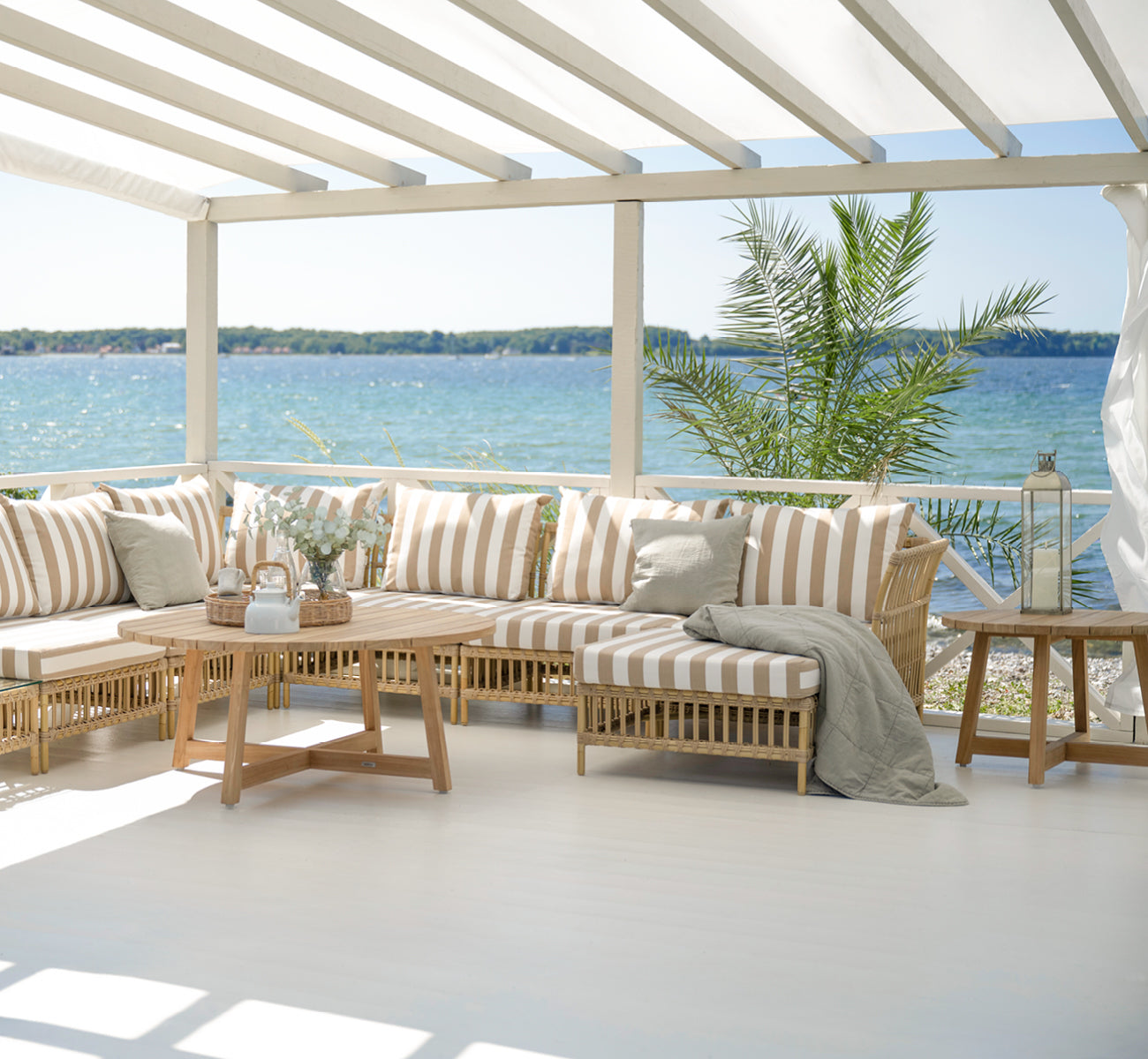 Esta es una imagen de estilo de vida de Victor Teak Lounge Table Ø75 cm en teak outdoor. Colocada en una terraza junto al mar entre divanes con cojines a rayas.