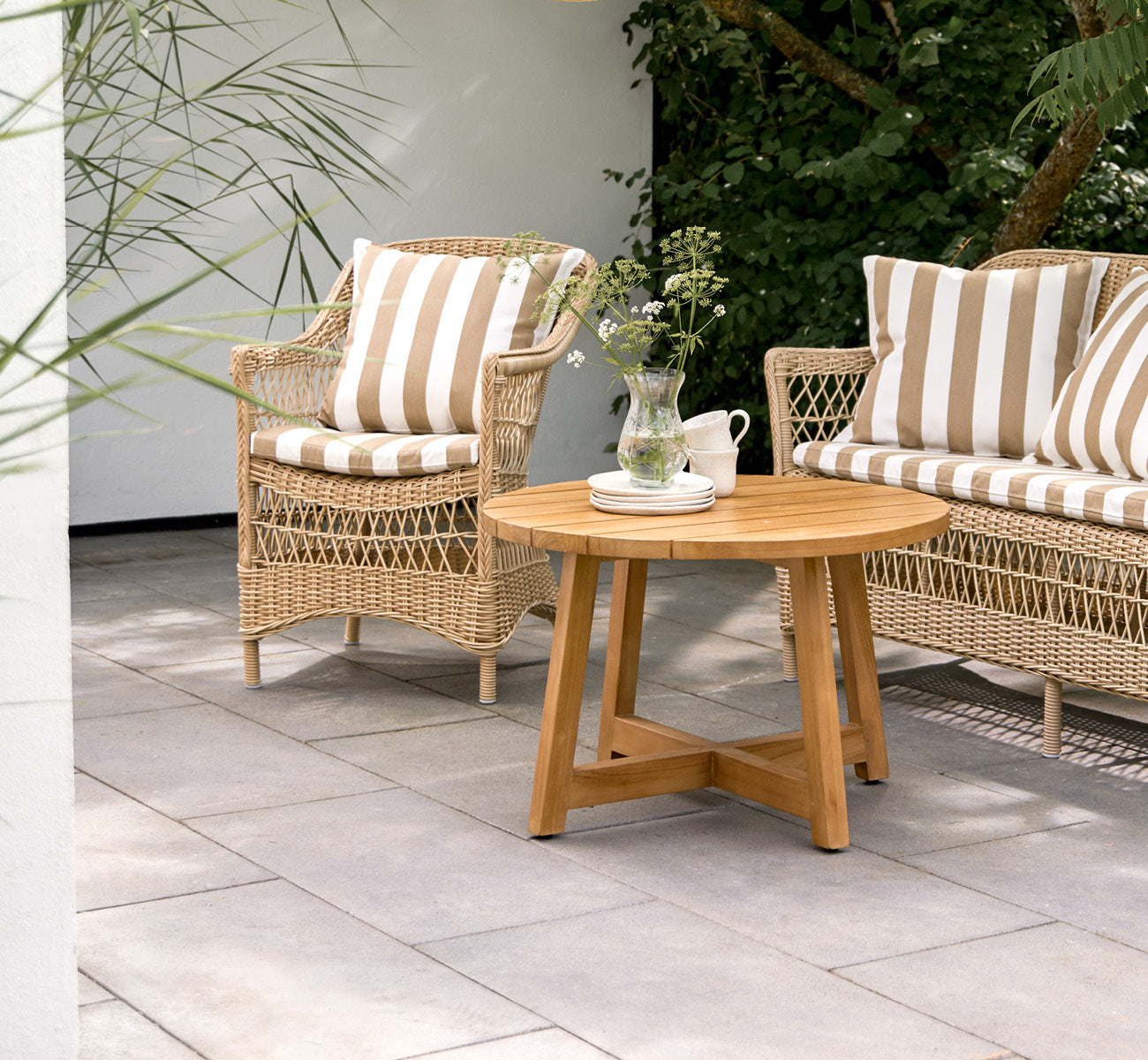Esta es una imagen de estilo de vida de Victor Teak Lounge Table Ø75 cm en teak outdoor. Colocada en una terraza junto al mar entre divanes con cojines a rayas.