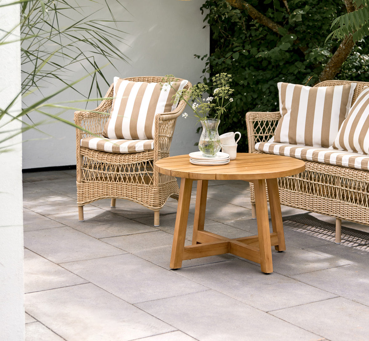Esta es una imagen de estilo de vida de Victor Teak Lounge Table Ø75 cm en teak outdoor. Colocada en una terraza junto al mar entre divanes con cojines a rayas.