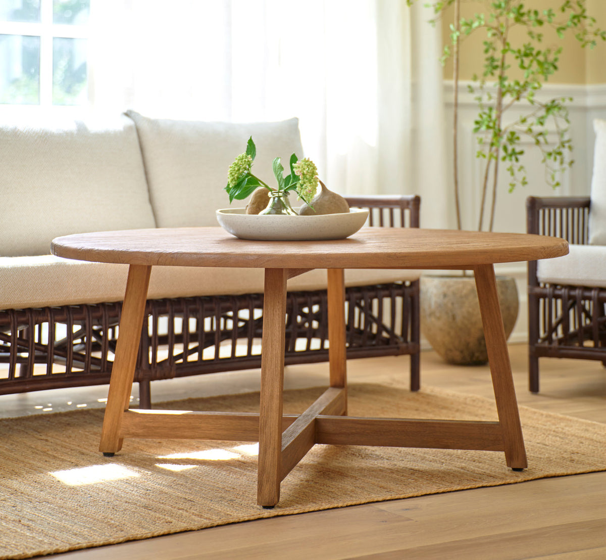 Esta es una imagen de estilo de vida de Aksel mesa de centro Ø120 cm en teak indoor. Delante de un sofá de ratán en un salón soleado.