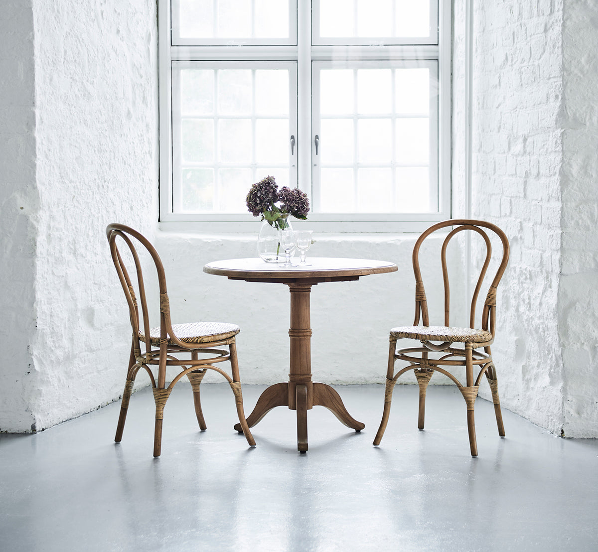 Esta es una imagen de estilo de vida de Michel Teak Dining Table Ø80 cm en teak indoor. Con dos sillas y flores en un interior luminoso de ladrillo blanco.