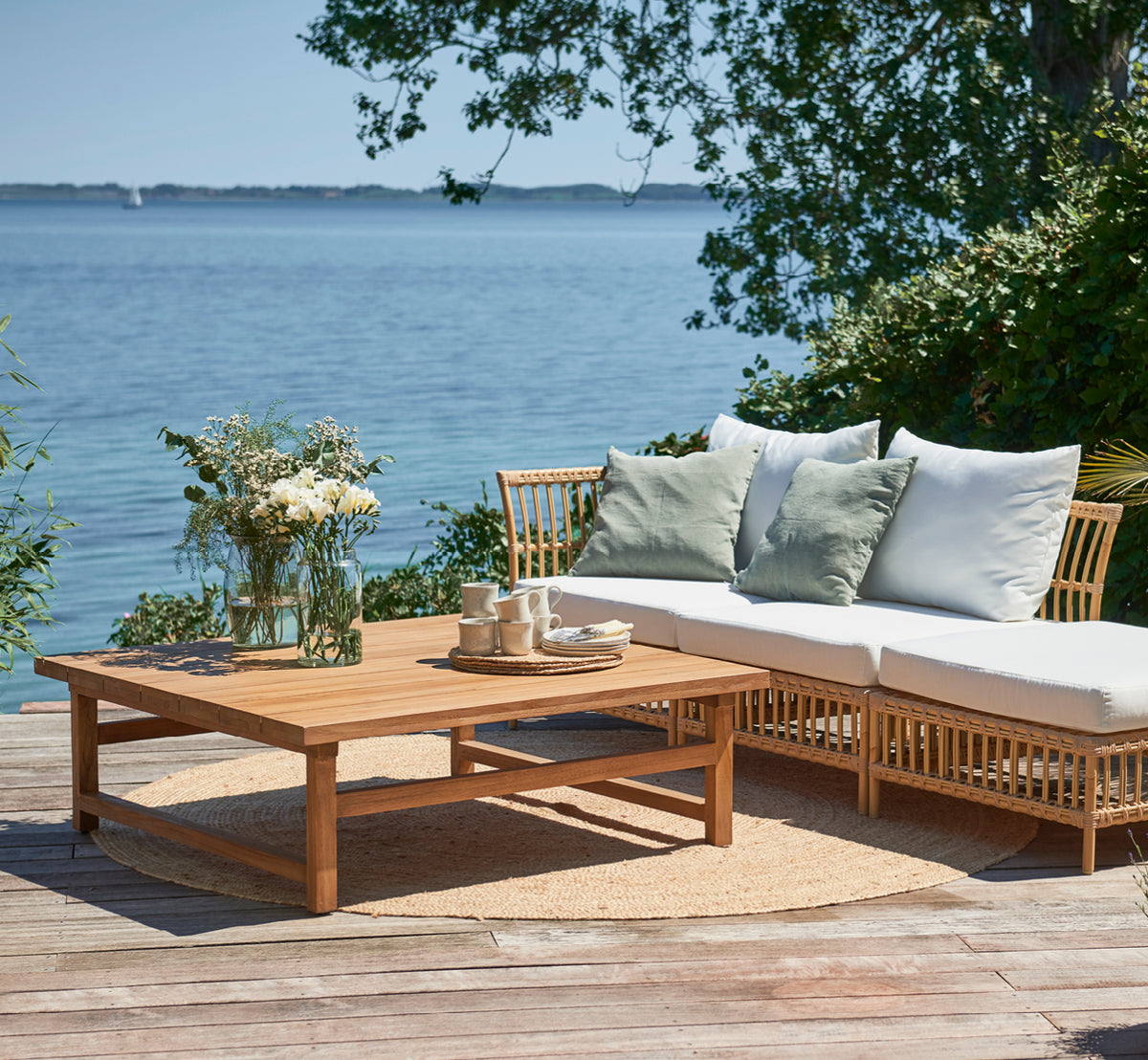 Esta es una imagen de estilo de vida de Julian Teak Lounge Table 120x120 cm en teak outdoor. En una terraza junto al mar con un sofá lounge, cojines y un juego de té.