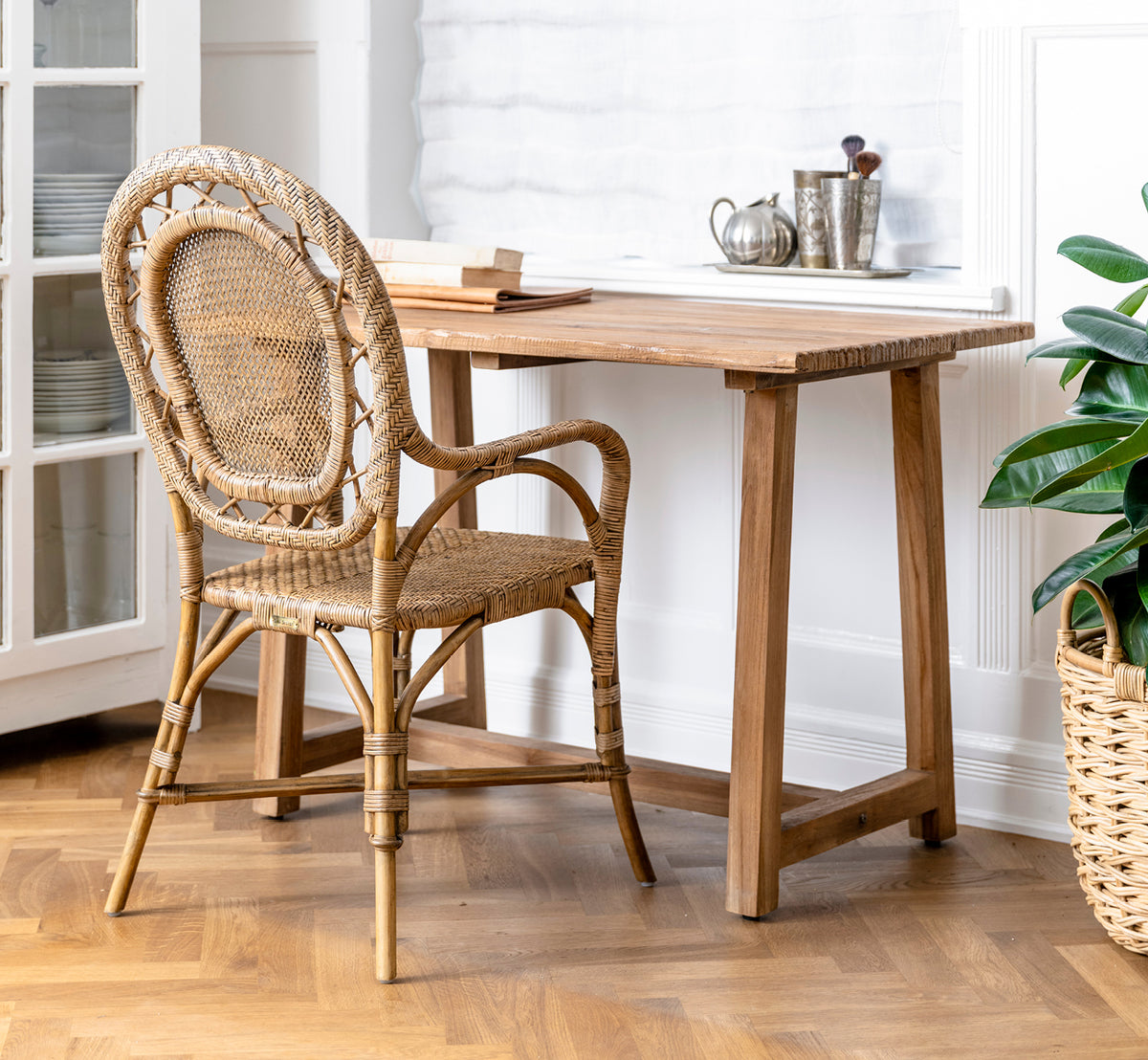 Esta es una imagen de estilo de vida de Lucas Teak escritorio en teak indoor. Combinado con una silla de ratán y planta verde en un interior luminoso con suelo en espiga.