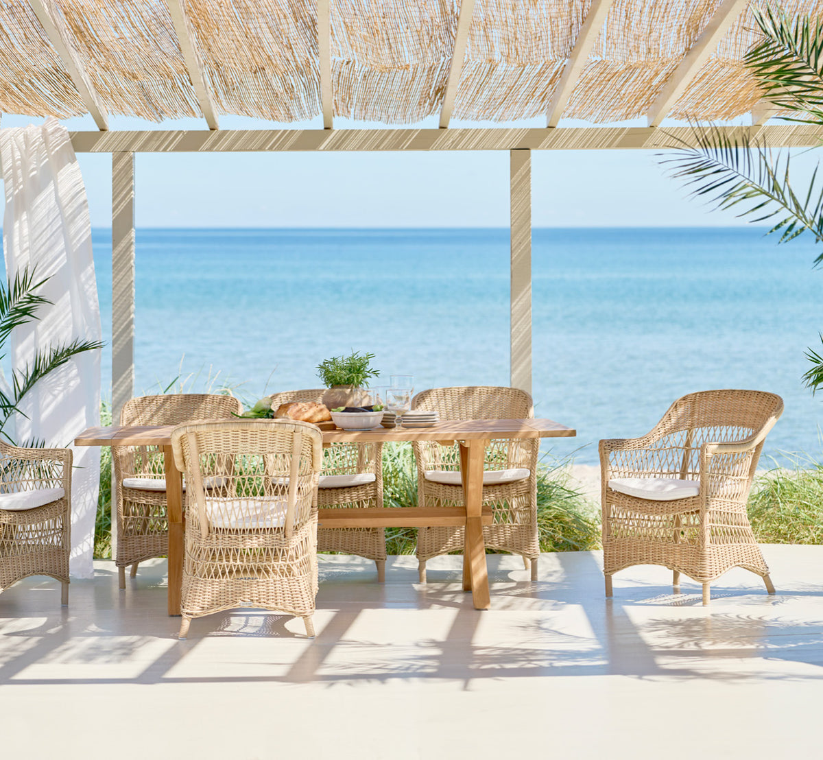 Esta es una imagen de estilo de vida de Colonial mesa 200 en outdoor Teak, en una terraza junto a la playa con cubierta de madera blanca y vistas al.