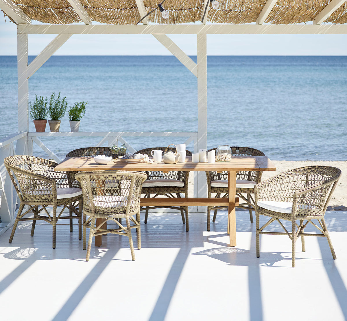 Esta es una imagen de estilo de vida de Colonial mesa 200 en outdoor Teak, en una terraza junto a la playa con cubierta de madera blanca y vistas al.