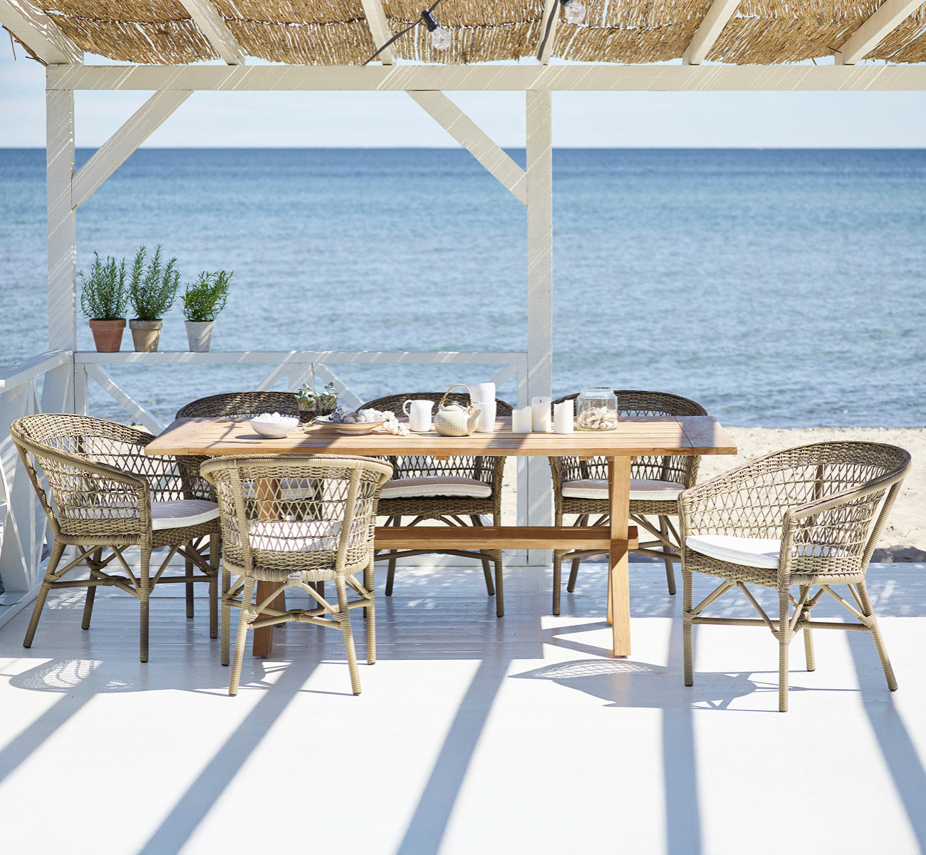 Esta es una imagen de estilo de vida de Colonial mesa 200 en outdoor Teak, en una terraza junto a la playa con cubierta de madera blanca y vistas al.