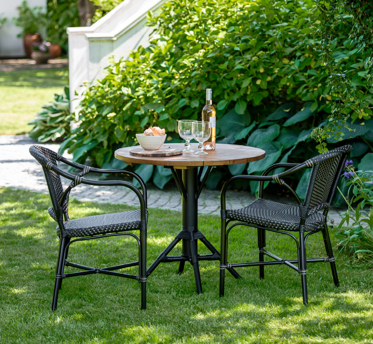 Esta es una imagen de estilo de vida de Nicole Cafe Table i black aluminium frame black ArtFibre. En una terraza de piedra con vegetación de jardín.
