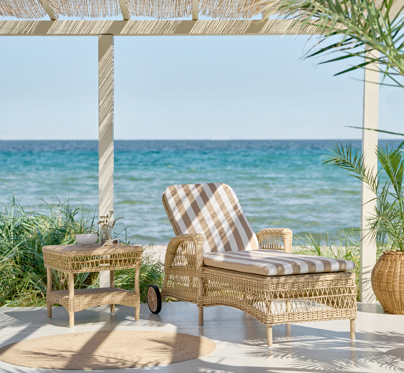 Esta es una imagen de estilo de vida de Susy mesa auxiliar en natural ArtFibre Aluminium, en una terraza costera con vistas al mar, gramíneas de.