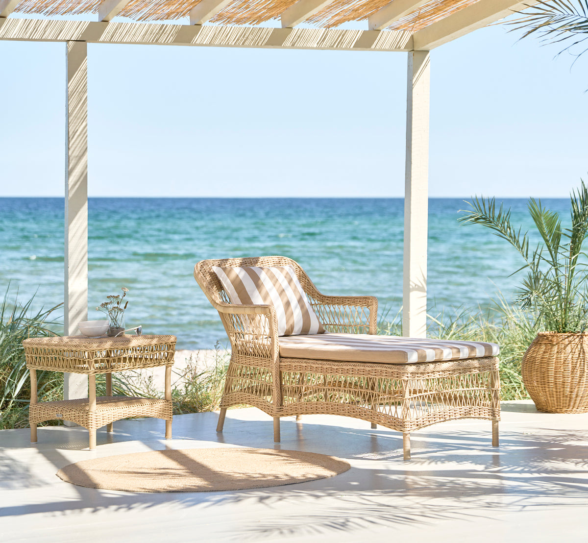 Esta es una imagen de estilo de vida de Olivia Exterior Sunbed i natural artfibre aluminium. En un jardín exuberante con cojín y manta.