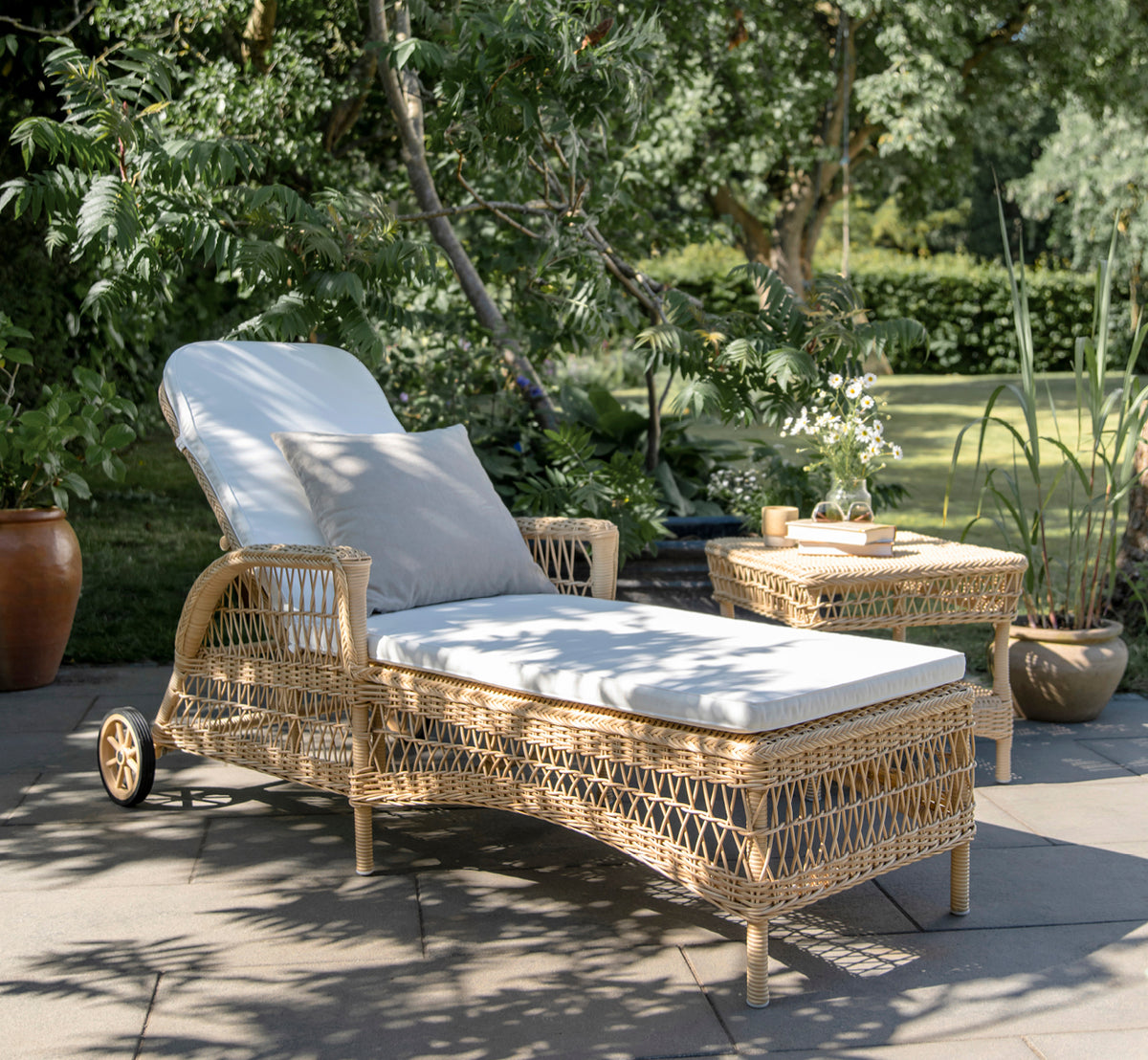 Esta es una imagen de estilo de vida de Daisy tumbona en natural ArtFibre Aluminium, en una terraza de jardín rodeada de frondosos árboles y plantas.