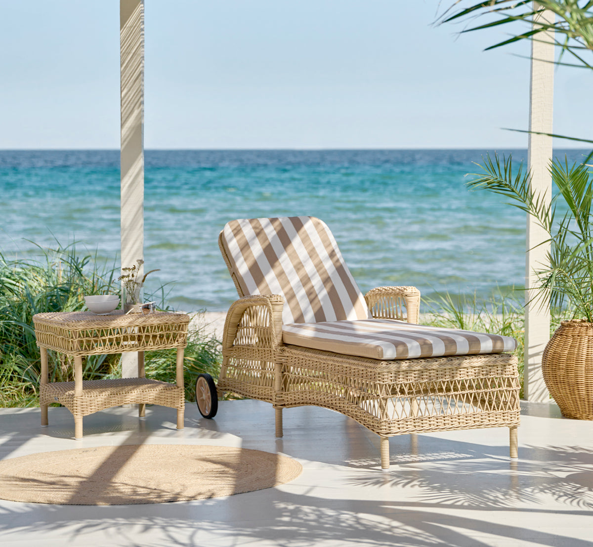 Esta es una imagen de estilo de vida de Daisy tumbona en natural ArtFibre Aluminium, en una terraza de jardín rodeada de frondosos árboles y plantas.