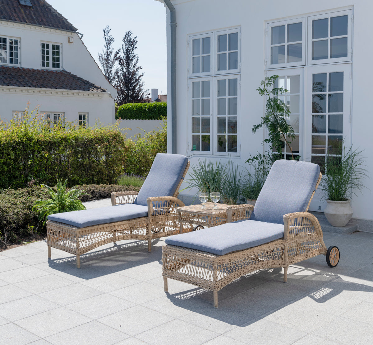 Esta es una imagen de estilo de vida de Daisy tumbona en natural ArtFibre Aluminium, en una terraza de jardín rodeada de frondosos árboles y plantas.