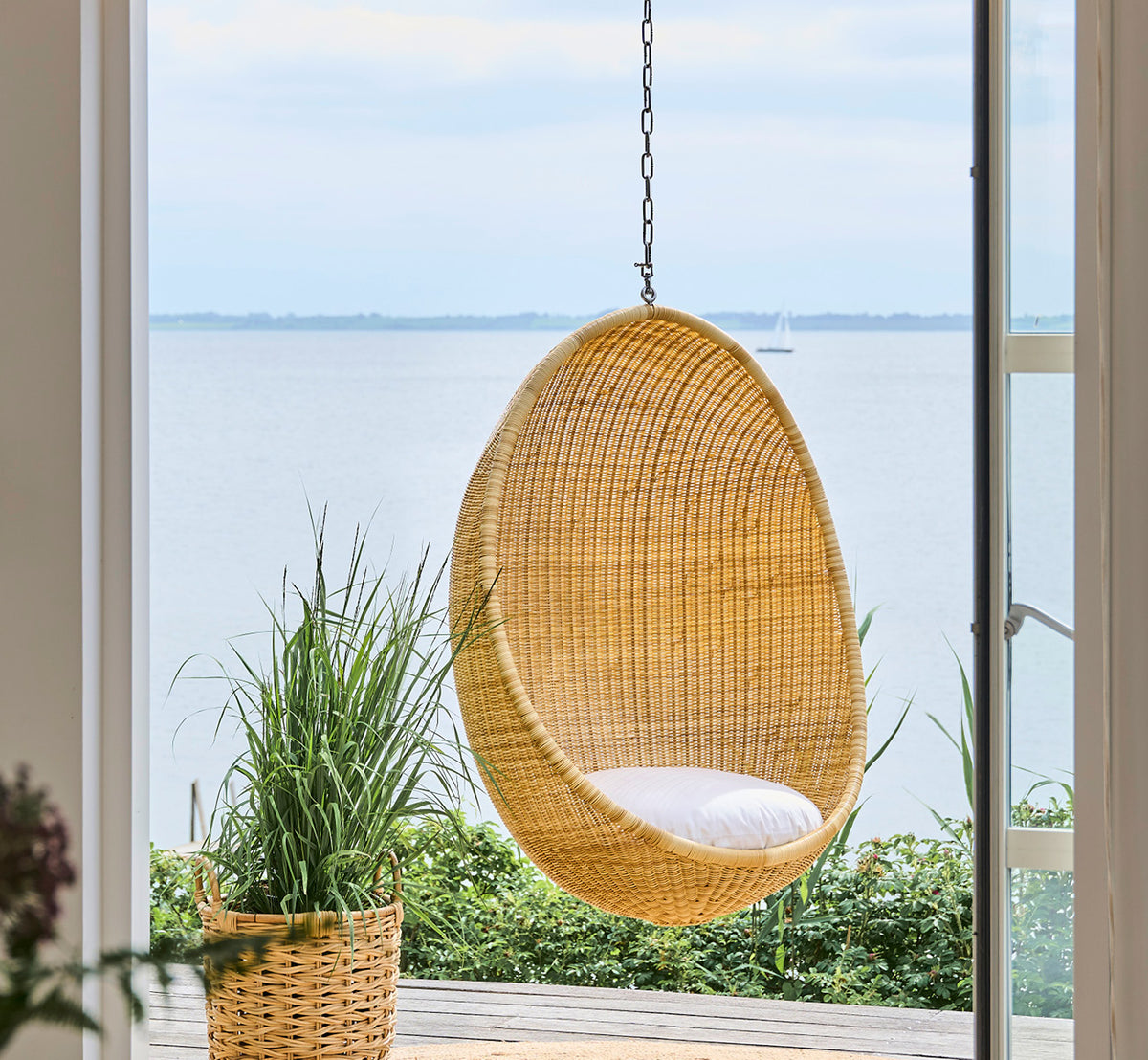 Esta es una imagen de estilo de vida de Chain huevo colgante en metal, colgado junto a una puerta de cristal con vistas al agua y plantas verdes.
