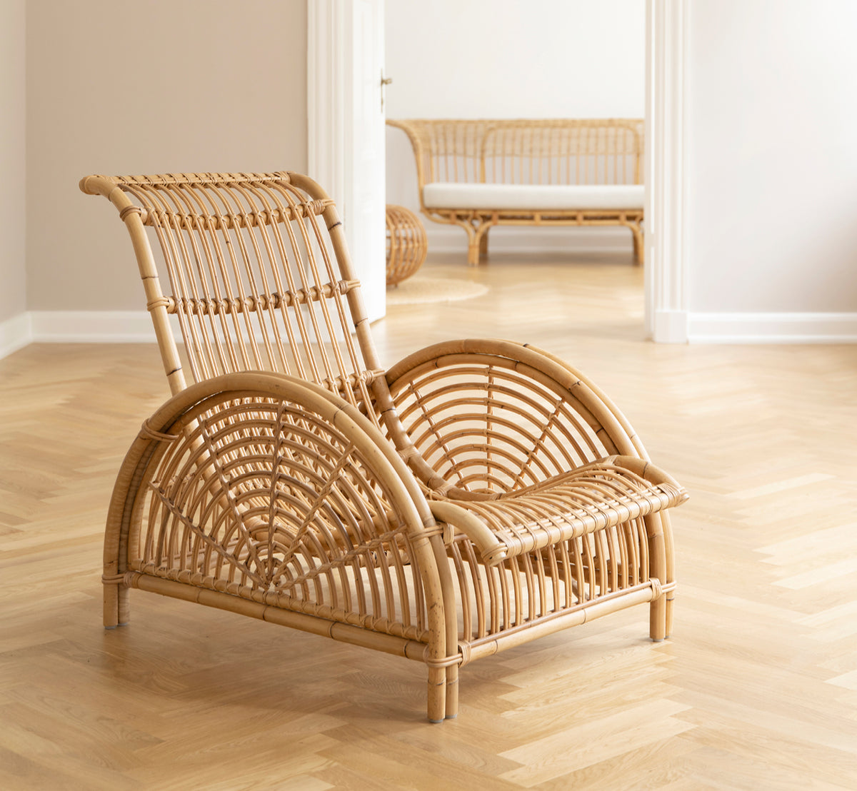 Esta es una imagen de estilo de vida de Paris sillón lounge en polished natural rattan. Colocada sobre un suelo de parqué en espiga junto a un banco a juego.