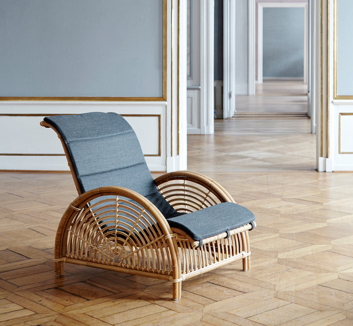 Esta es una imagen de estilo de vida de Paris sillón lounge en polished natural rattan. Colocada sobre un suelo de parqué en espiga junto a un banco a juego.