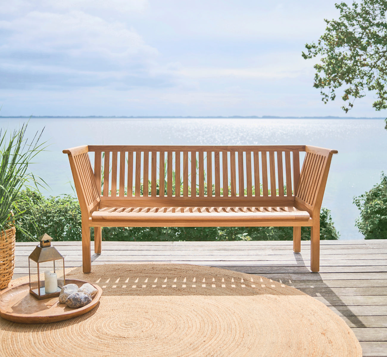 Esta es una imagen de estilo de vida de St Catherine banco de teca en teak outdoor. Colocado en una cubierta de madera con vistas al mar y una alfombra redonda de yute.