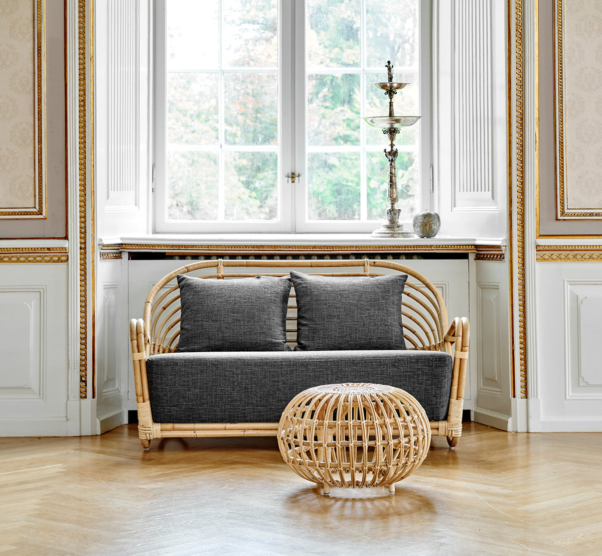 Esta es una imagen de estilo de vida de Charlottenborg sofá en polished Natural Rattan, en un elegante interior clásico con elaboradas molduras.