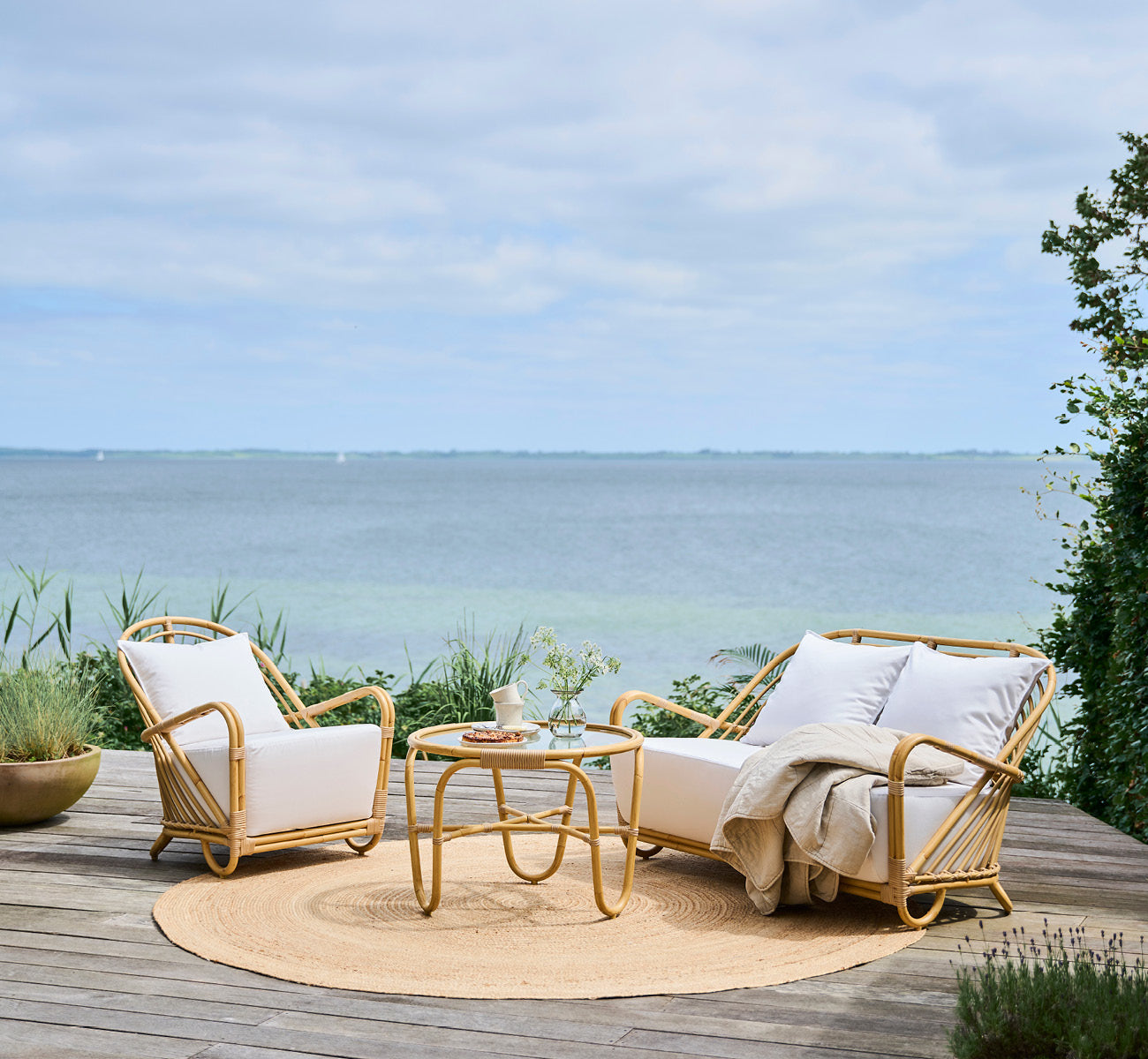 Esta es una imagen de estilo de vida de Charlottenborg sillón lounge exterior en natural Aluminium ArtFibre, en una terraza de madera al aire libre.
