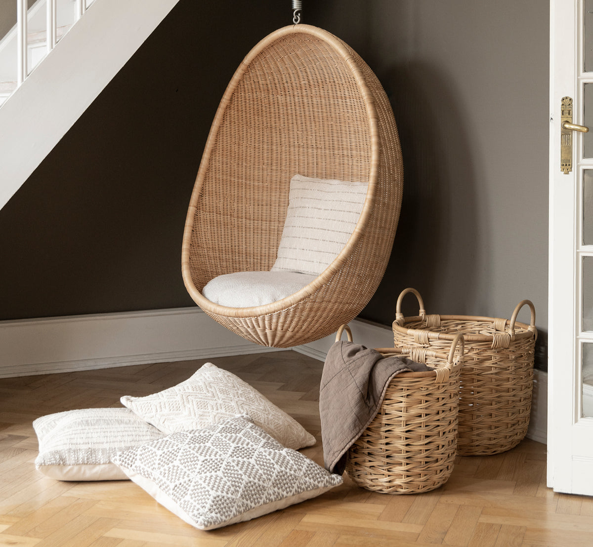 Esta es una imagen de estilo de vida de Sarah juego de cestas en natural rattan. Colocada junto a una silla colgante tipo pod y cojines estampados sobre parqué.