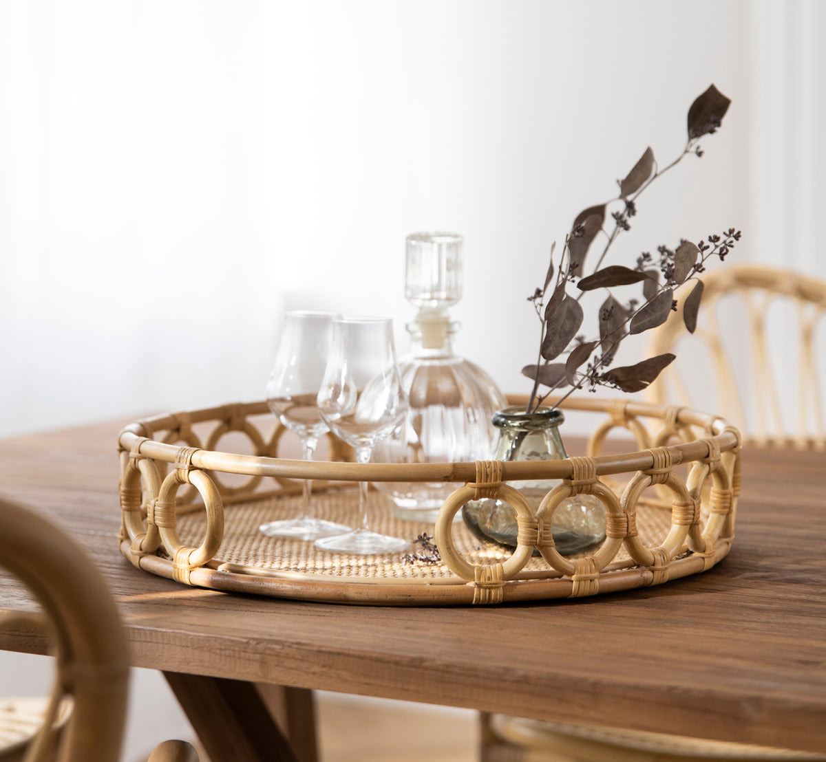 Esta es una imagen de estilo de vida de Mira Tray i natural rattan. Sobre una mesa de comedor con cristalería y eucalipto seco en una sala luminosa.