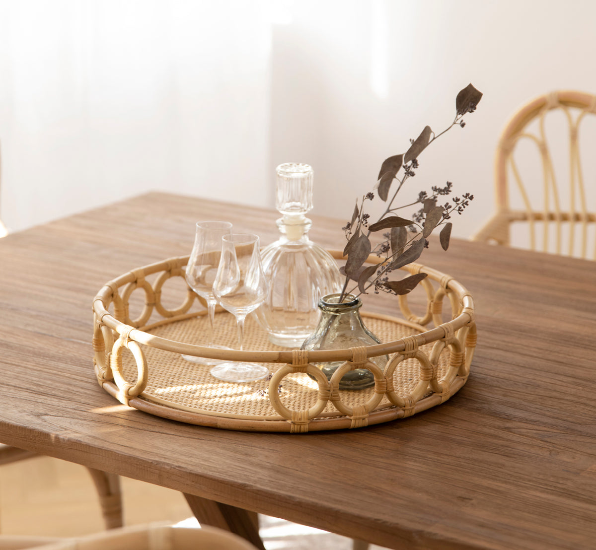 Esta es una imagen de estilo de vida de Mira Tray i natural rattan. Sobre una mesa de teca con copas de cristal y un jarrón con ramas secas.