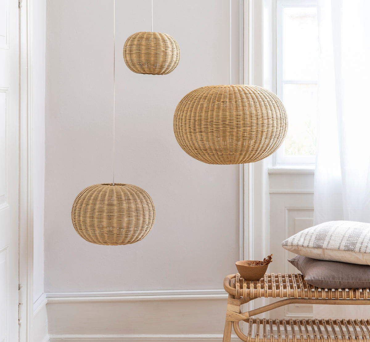 Esta es una imagen de estilo de vida de Tangelo Lampshade Ø37 cm | Small en natural rattan. Tres lámparas colgantes agrupadas sobre un banco de ratán en un interior luminoso.