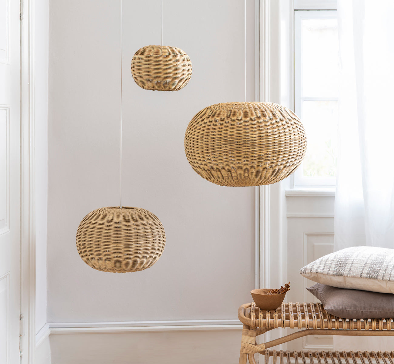 Esta es una imagen de estilo de vida de Tangelo Lampshade Ø49 cm | Medium en natural rattan. Tres lámparas colgantes contra una pared blanca sobre un banco de ratán.