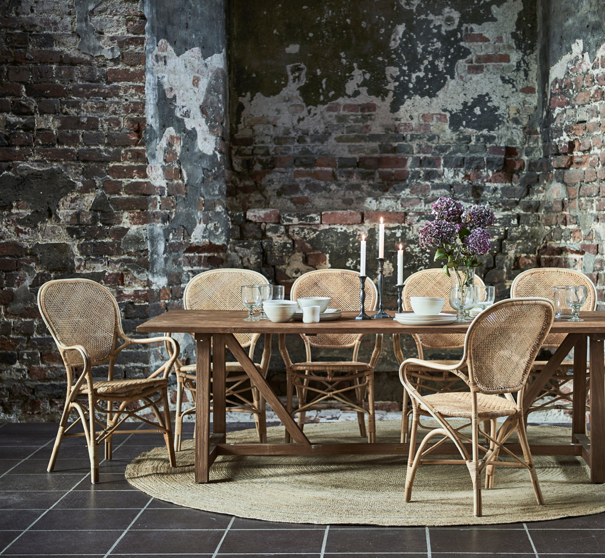 Esta es una imagen de estilo de vida de Jute Carpet Ø280 en natural jute. Bajo una mesa de comedor con sillas de ratán en un espacio rústico con paredes de ladrillo.