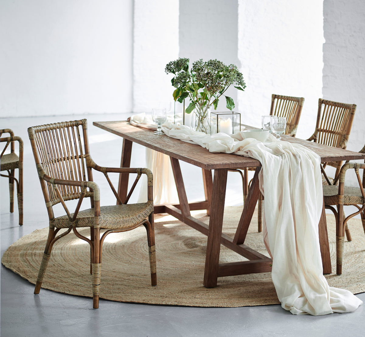 Esta es una imagen de estilo de vida de Jute Carpet Ø280 en grey jute. Bajo una mesa de comedor con sillas de ratán, lino drapeado y vegetación.