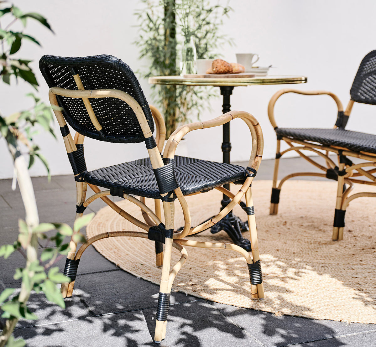 Esta es una imagen de estilo de vida de Bistro Dining Chair en skin on Rattan frame, en un ambiente de café al aire libre con una mesa de bistró.